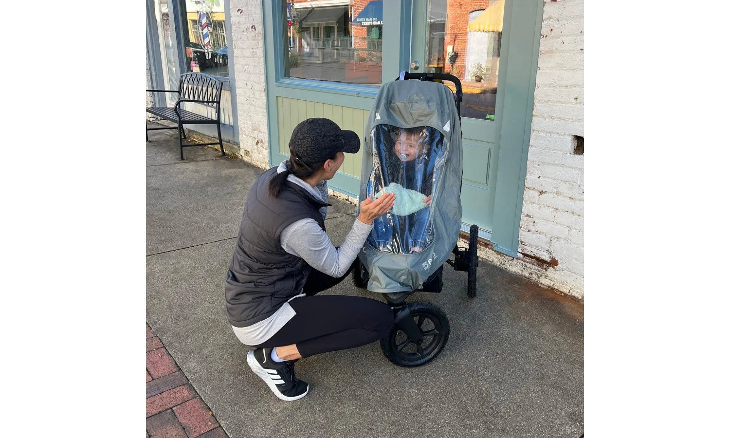 A woman kneeling beside a child in a jogging stroller. The stroller is covered by a weather shield.